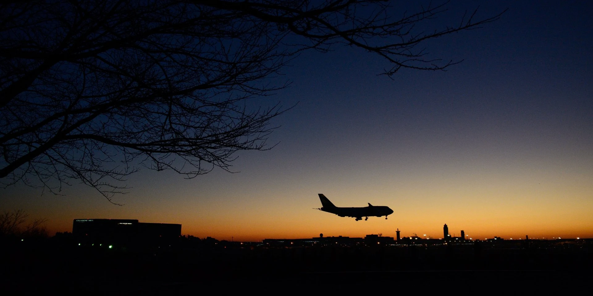 Narita Airport, wide-body aircraft on final approach against a deep orange-to-blue twilight gradient, bare winter tree branches top-left